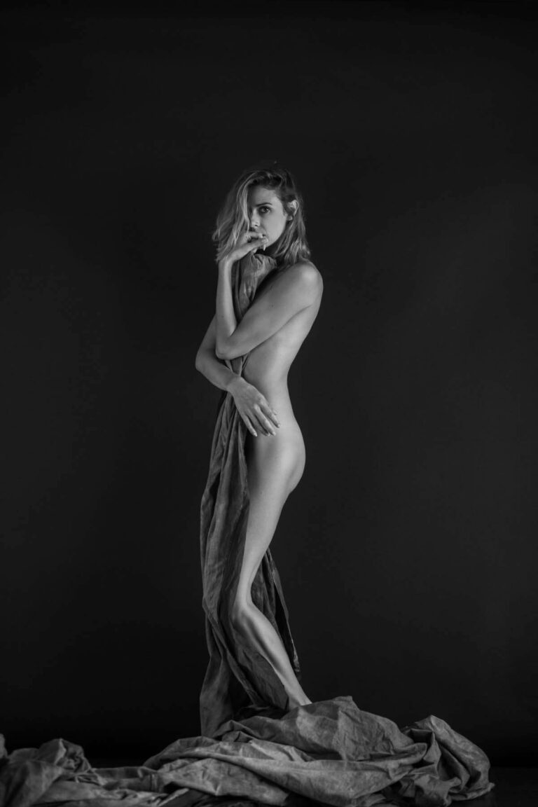 Une personne nue se tient de profil sur un fond sombre, partiellement recouverte d'un tissu drapé. Elle porte une main à son visage et a une expression contemplative. Le tissu est étalé sur le sol autour de ses pieds. L'image est en noir et blanc.