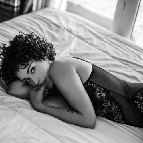 Une personne aux cheveux bouclés est allongée sur le côté d'un lit, vêtue d'un body en dentelle. Elle repose sa tête sur un bras et regarde l'appareil photo, un doigt près de ses lèvres. La photo est en noir et blanc, avec une lumière naturelle provenant d'une fenêtre.