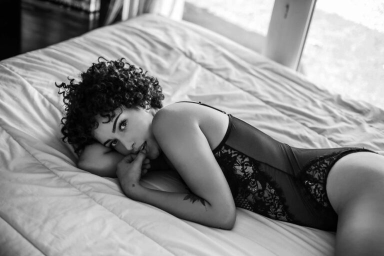 Une personne aux cheveux bouclés est allongée sur le côté d'un lit, vêtue d'un body en dentelle. Elle repose sa tête sur un bras et regarde l'appareil photo, un doigt près de ses lèvres. La photo est en noir et blanc, avec une lumière naturelle provenant d'une fenêtre.