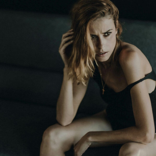 Une femme aux cheveux châtain clair est assise sur un canapé bleu foncé, vêtue d'une tenue noire dont l'une des bretelles glisse sur son épaule. Elle regarde sur le côté avec une expression sérieuse, sa main droite touchant ses cheveux, et un collier en or est visible autour de son cou.