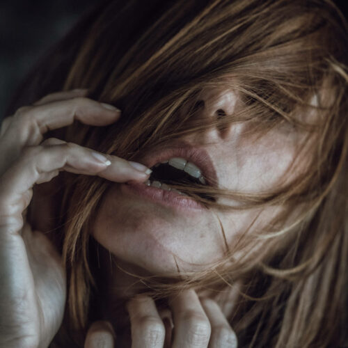 Gros plan d'une personne à la peau claire et aux cheveux bruns mi-longs couvrant la majeure partie de son visage. Sa bouche est légèrement ouverte et ses doigts touchent délicatement ses lèvres et son menton, créant ainsi une atmosphère intime et morose. L'arrière-plan est légèrement flou.