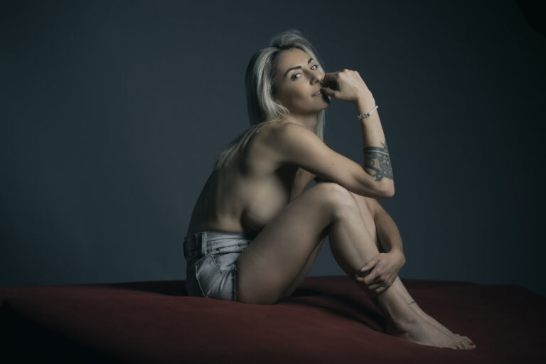 Une personne aux seins nus, aux longs cheveux blonds platine, ne portant qu'un short en jean clair, est assise sur une surface rouge sur un fond sombre. Elle a un tatouage sur le bras et pose son menton sur sa main, regardant pensivement la caméra.