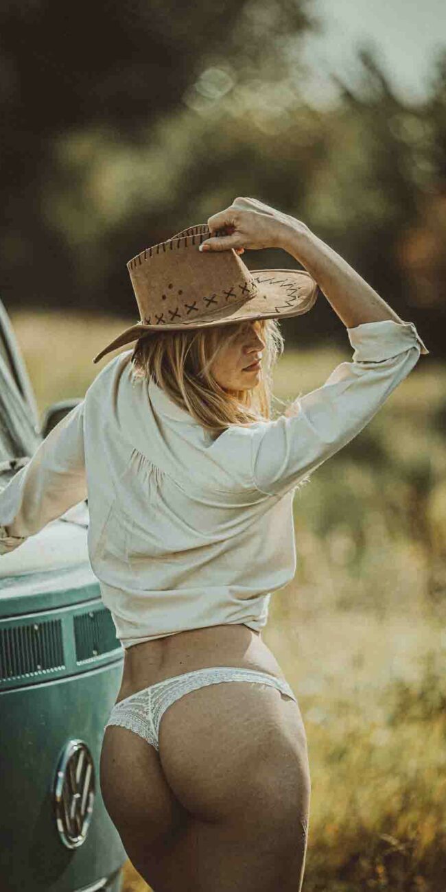 Une femme vêtue d'une chemise blanche, d'un slip blanc en dentelle et d'un chapeau de cow-boy marron se tient à l'extérieur, à côté d'une camionnette Volkswagen de couleur sarcelle. Elle regarde au loin, par-dessus son épaule, sa main tenant le bord de son chapeau. L'arrière-plan est herbeux, avec des arbres flous.