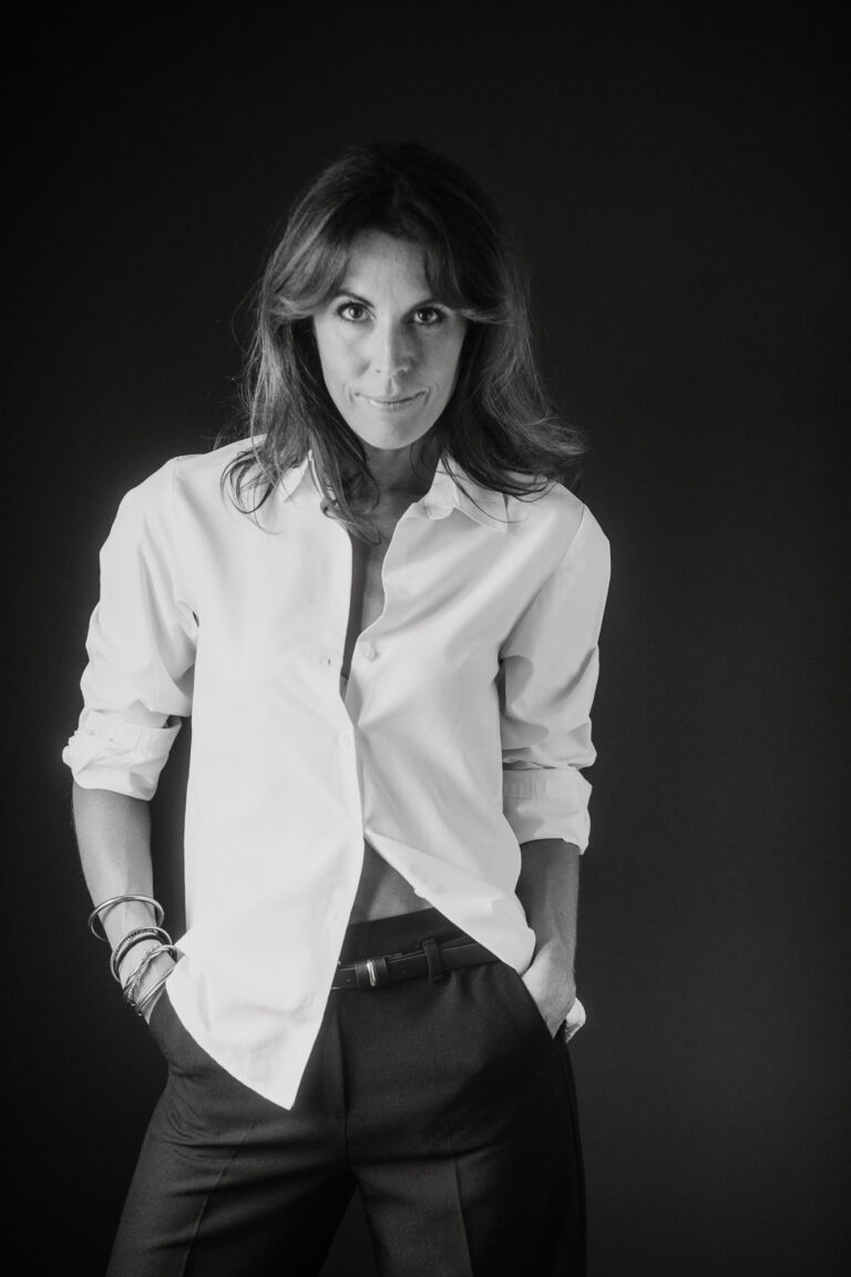 Portrait en noir et blanc réalisé par un photographe : une femme se tient debout sur un fond sombre, vêtue d'une chemise blanche boutonnée aux manches retroussées et d'un pantalon sombre, ses cheveux ondulés encadrant une pose confiante, semblable à celle d'un mannequin, et un sourire subtil.