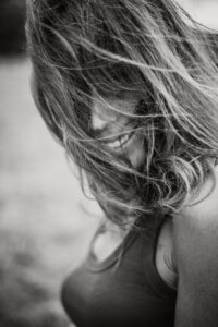 Photo en noir et blanc d'une personne ressemblant à un mannequin, dont les cheveux longs couvrent la majeure partie du visage, et qui regarde vers le bas. Le sujet, qui rappelle le portrait d'un livre de photographie, porte un haut sans manches sur un fond extérieur légèrement flou.