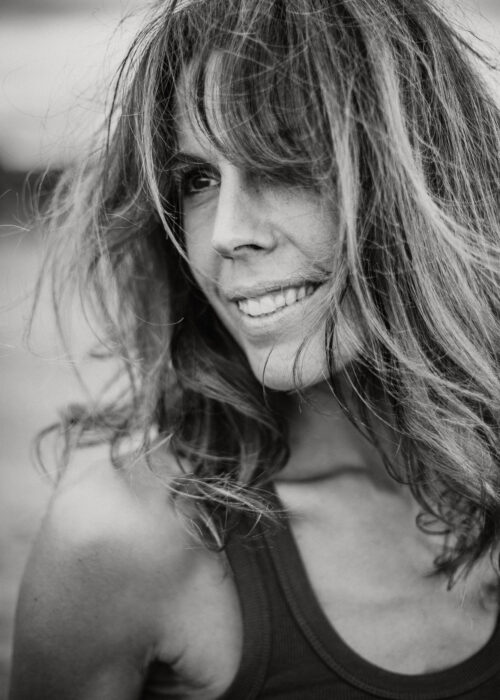 Photo en noir et blanc d'une femme dont les longs cheveux ondulés couvrent partiellement le visage. Elle sourit doucement, comme un mannequin posant pour un livre de photos. L'arrière-plan flou permet de se concentrer sur son visage expressif.