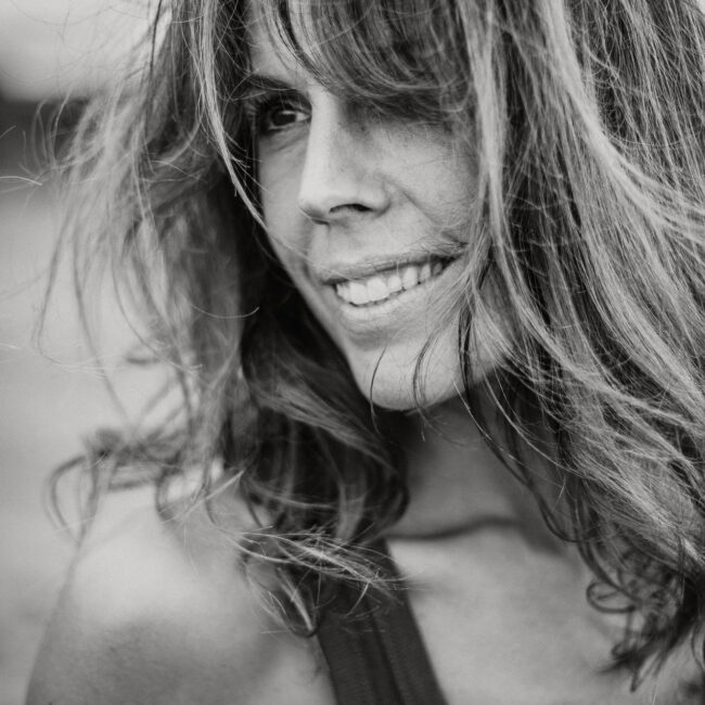 Photo en noir et blanc d'une femme dont les longs cheveux ondulés couvrent partiellement le visage. Elle sourit doucement, comme un mannequin posant pour un livre de photos. L'arrière-plan flou permet de se concentrer sur son visage expressif.