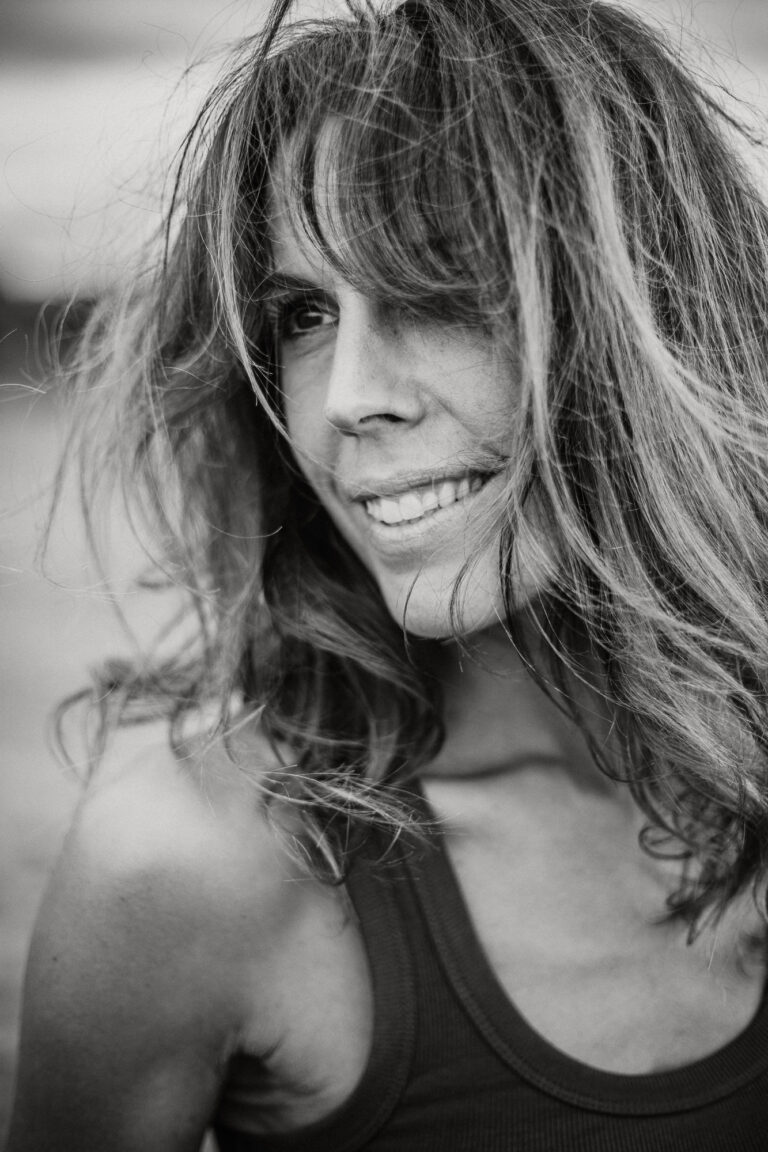 Photo en noir et blanc d'une femme dont les longs cheveux ondulés couvrent partiellement le visage. Elle sourit doucement, comme un mannequin posant pour un livre de photos. L'arrière-plan flou permet de se concentrer sur son visage expressif.