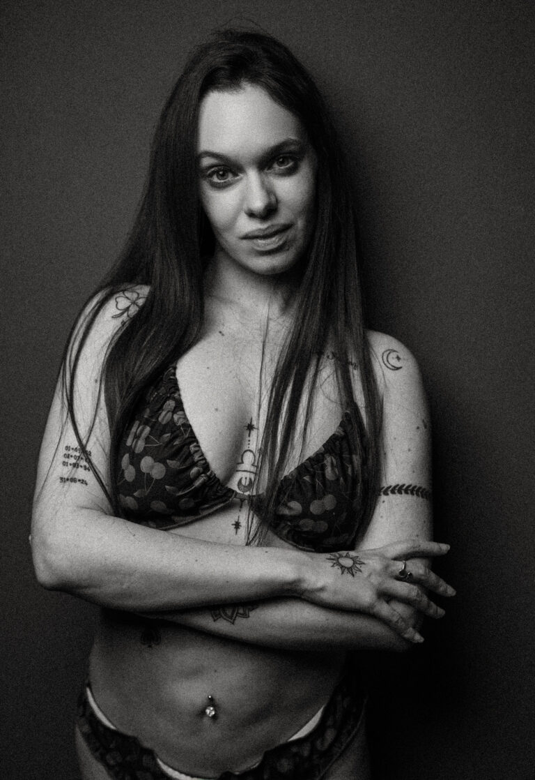 Une femme aux cheveux longs, portant un haut et un bas de bikini à motifs, se tient debout sur un fond uni, les bras croisés. Elle porte de nombreux tatouages visibles sur les bras, la poitrine et l'abdomen, ainsi qu'un piercing au nombril. L'image est en noir et blanc.