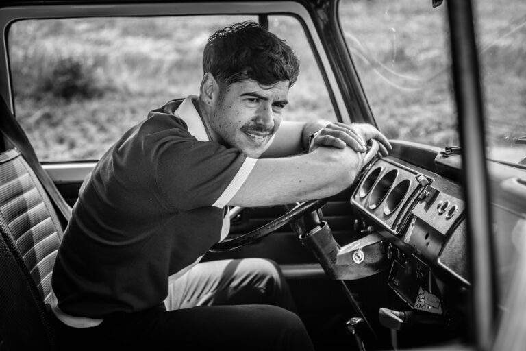 Photographie en noir et blanc d'un homme souriant appuyé sur le tableau de bord d'une voiture ancienne